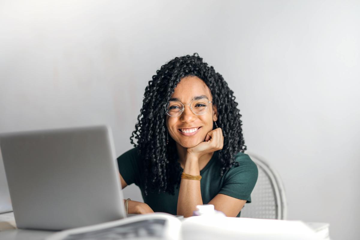 A young woman with curly black hair and tan skin look at the camera smiles while working on her laptop computer. 